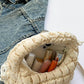 Quilted puffy crossbody bag with removable strap, open with cosmetics inside, next to a denim jacket.