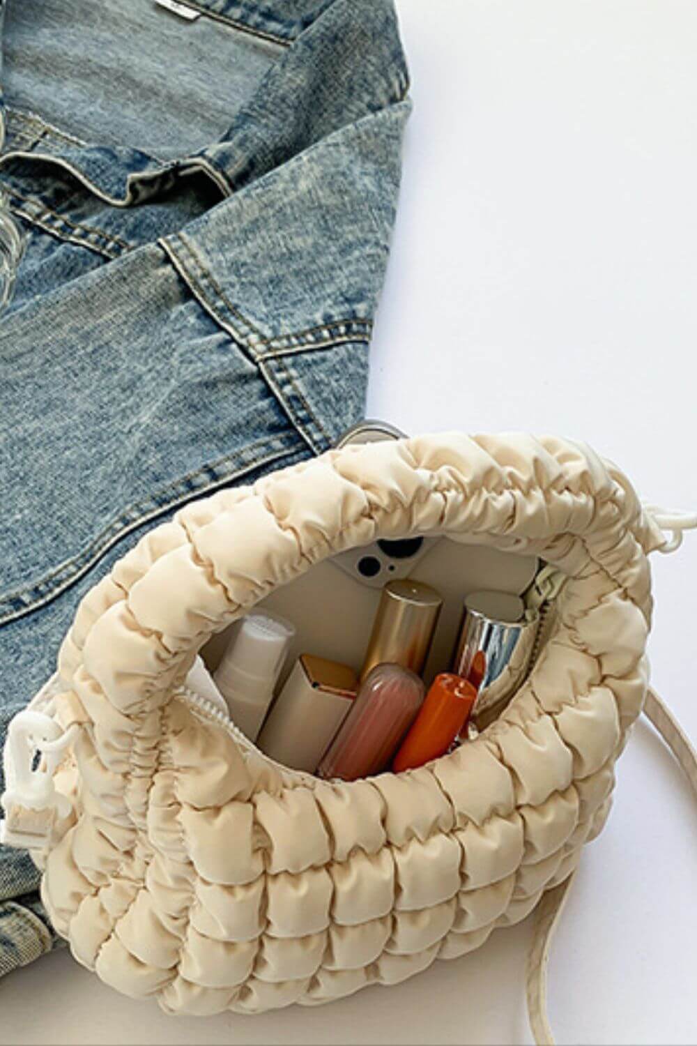 Quilted puffy crossbody bag with removable strap, open with cosmetics inside, next to a denim jacket.