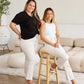 Two women in stylish white raw hem jeans and casual tops posing in a chic, modern living room setting.