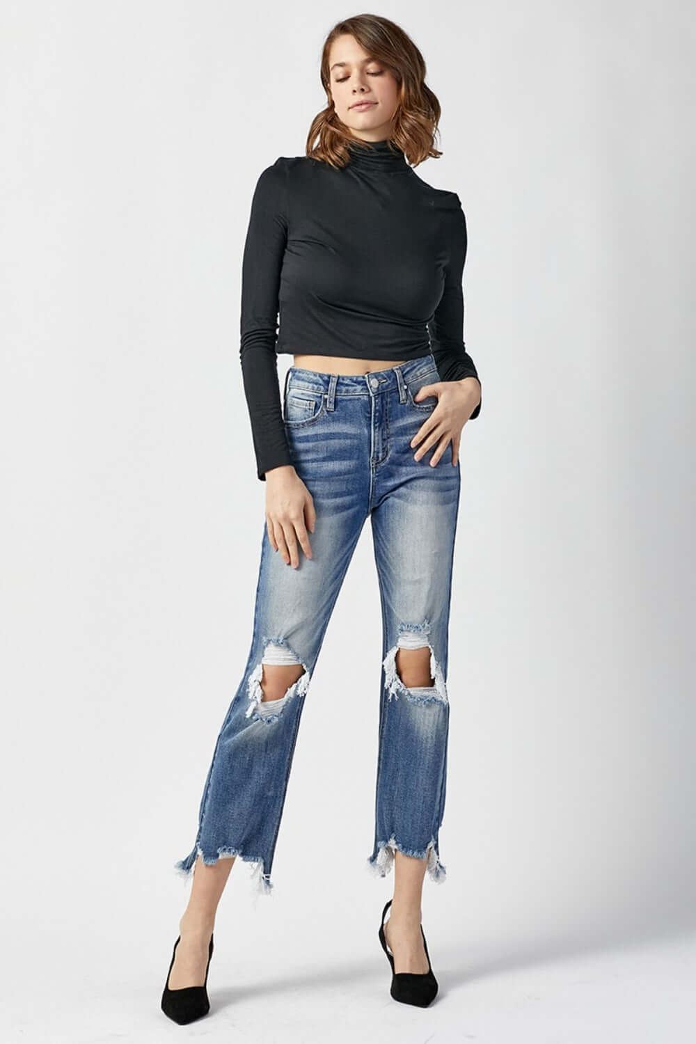 Woman wearing high waist distressed frayed hem cropped straight Risen Jeans styled with black heels and a fitted black top.