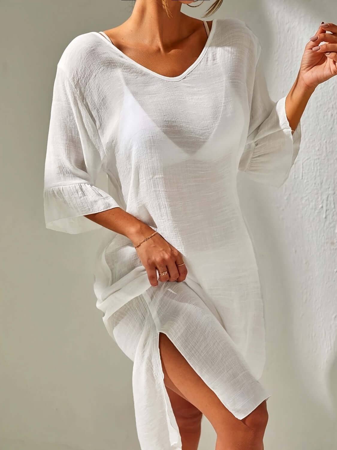 Woman wearing a white slit v-neck flounce sleeve cover-up, showcasing sheer and backless design with tied detail.