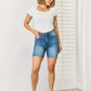 Woman wearing Tummy Control Double Button Bermuda Denim Shorts from Judy Blue Jeans, styled with a white top and casual shoes