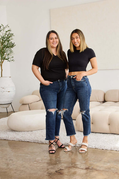 Two women wearing Tummy Control Distressed High Waist Raw Hem Jeans by RFM Jeans in living room setting