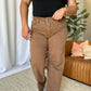 Woman wearing RFM Full Size High Rise Garment Dye Wide Leg Jeans in brown with a black top, standing in a stylish, modern living room.