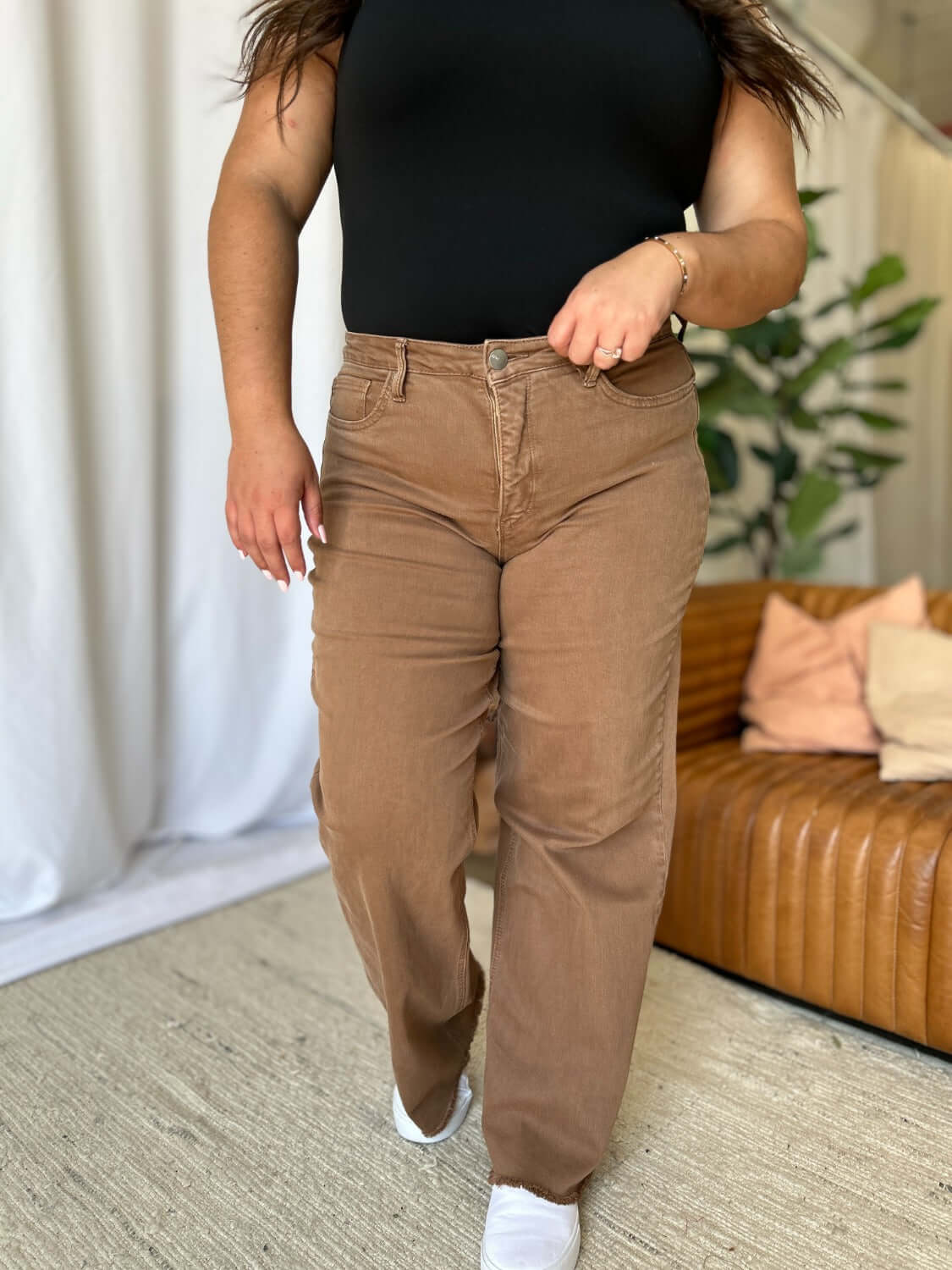 Woman wearing RFM Full Size High Rise Garment Dye Wide Leg Jeans in brown with a black top, standing in a stylish, modern living room.