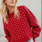 Woman wearing a red Bella Road sweater with a heart pattern and long sleeves, paired with jeans, showcasing a stylish fall look.