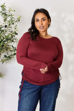 Plus-size model wearing a burgundy drawstring round neck long sleeve top and blue jeans, posing near a plant.