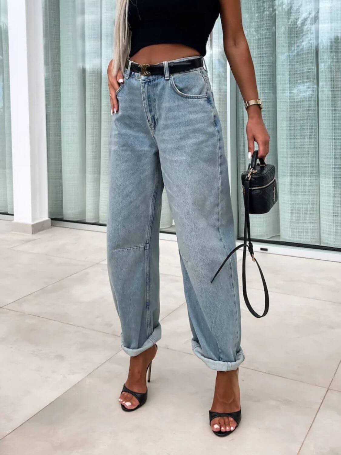 Woman wearing high waist wide leg jeans with a black crop top and heels, holding a black handbag in an indoor setting.