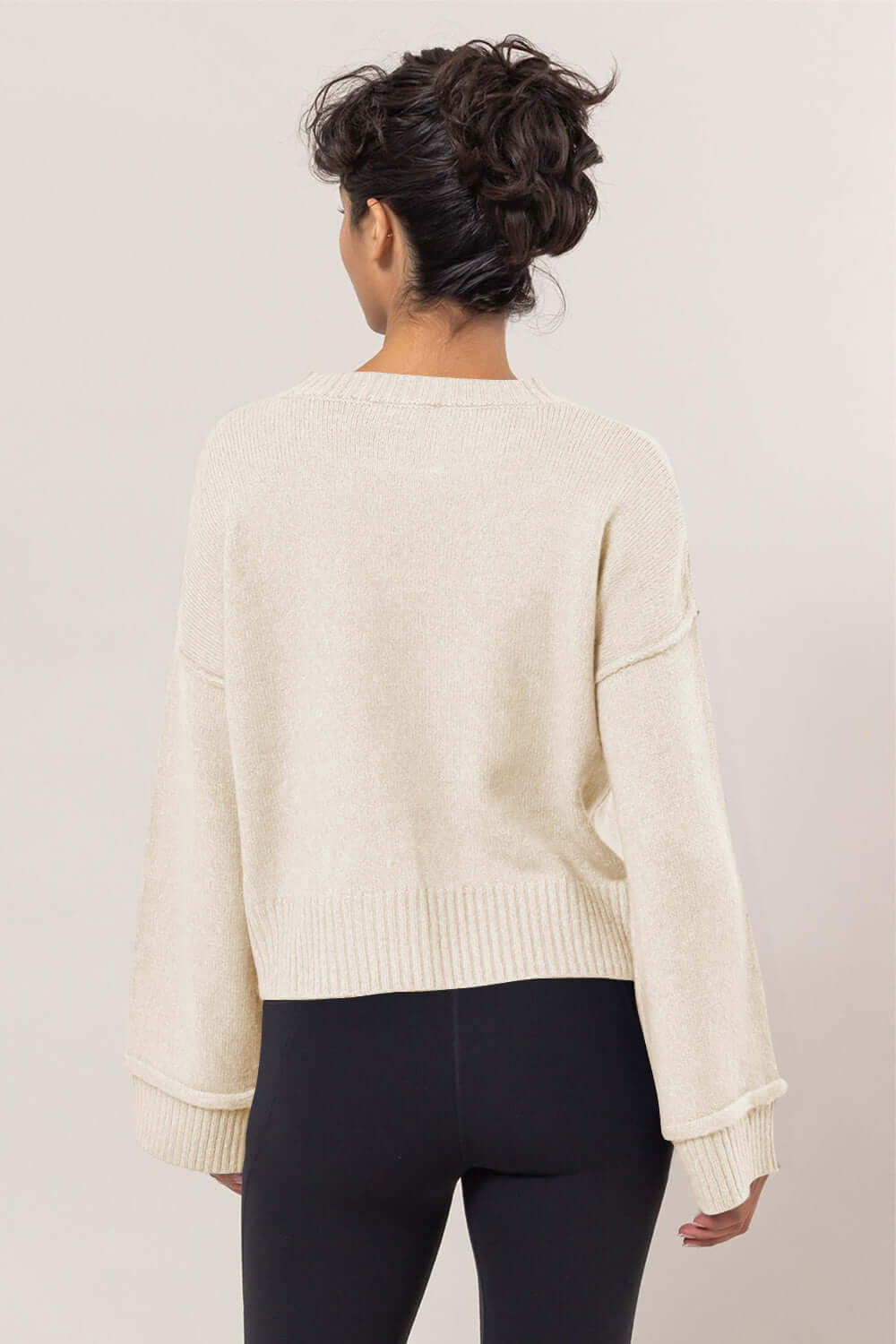 Back view of a woman in a cream dropped shoulder ribbed sweater paired with black leggings, showcasing a relaxed style.