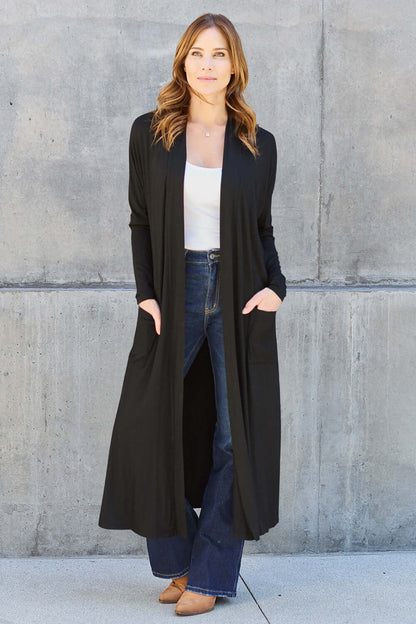 Woman wearing an open front long sleeve black cover up with pockets, over a white top and blue jeans, standing against a concrete wall