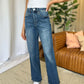 Woman wearing RFM Full Size High Rise Tummy Control Wide Leg Jeans paired with a white top, standing in a stylish living room.