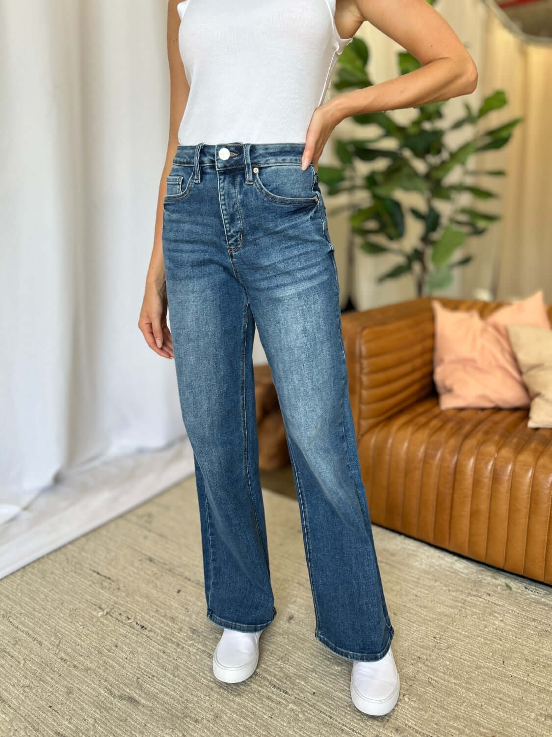 Woman wearing RFM Full Size High Rise Tummy Control Wide Leg Jeans paired with a white top, standing in a stylish living room.