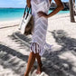 Woman in a white fringe cover-up walking on the beach, enjoying a sunny day by the turquoise sea.