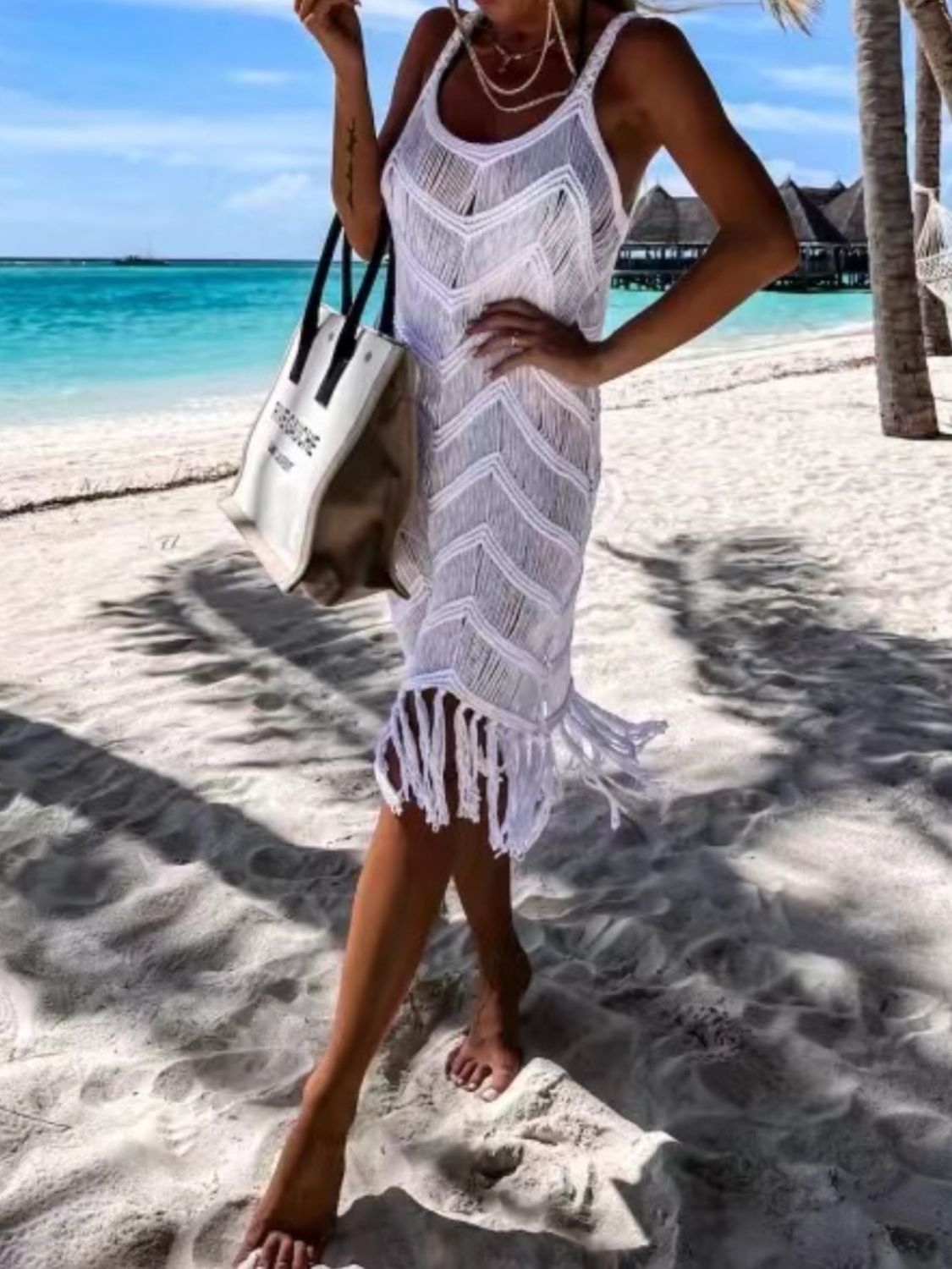 Woman in a white fringe cover-up walking on the beach, enjoying a sunny day by the turquoise sea.