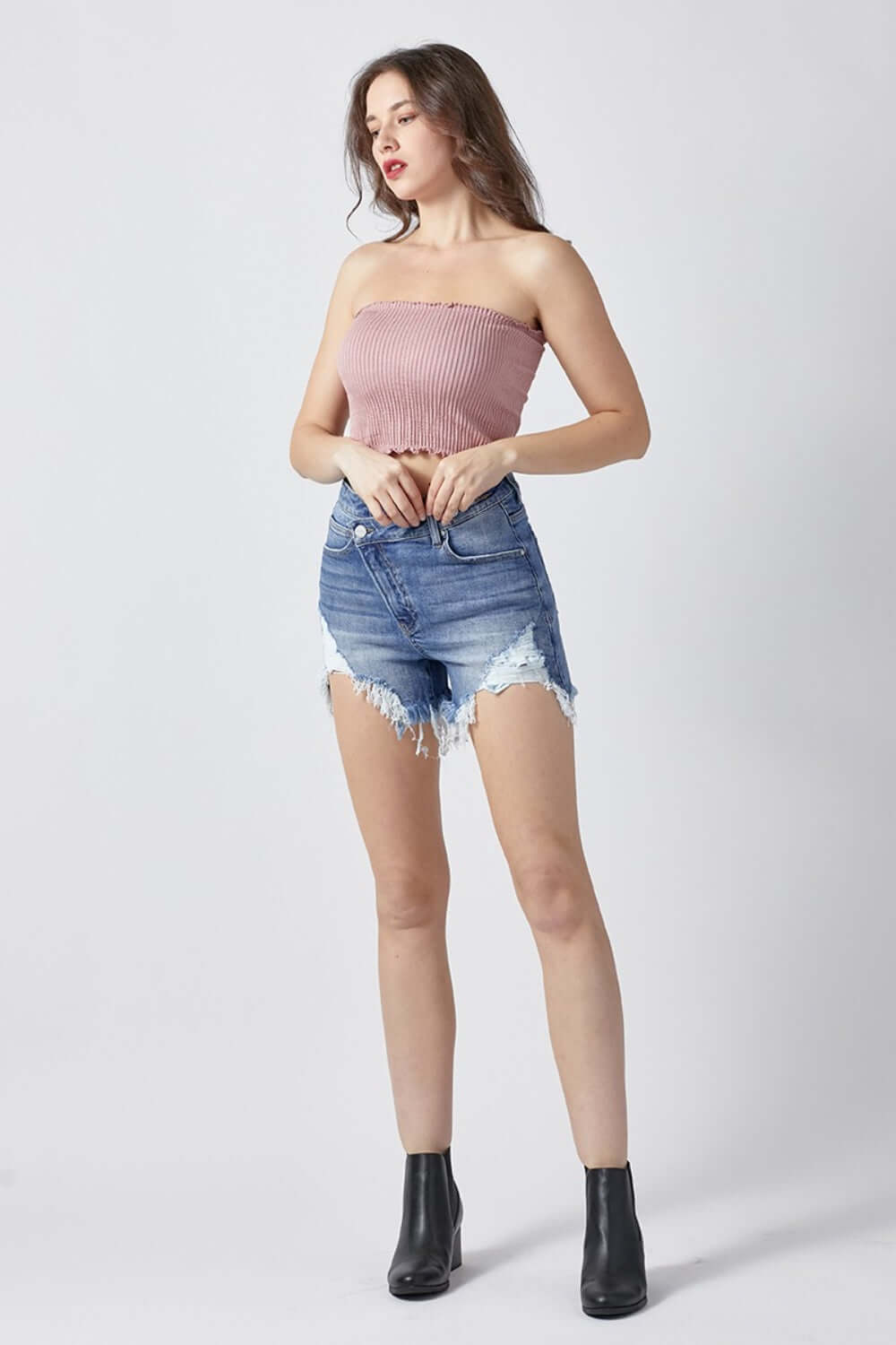 Woman wearing raw hem asymmetrical waist denim shorts by Risen Jeans with a pink strapless top and black ankle boots