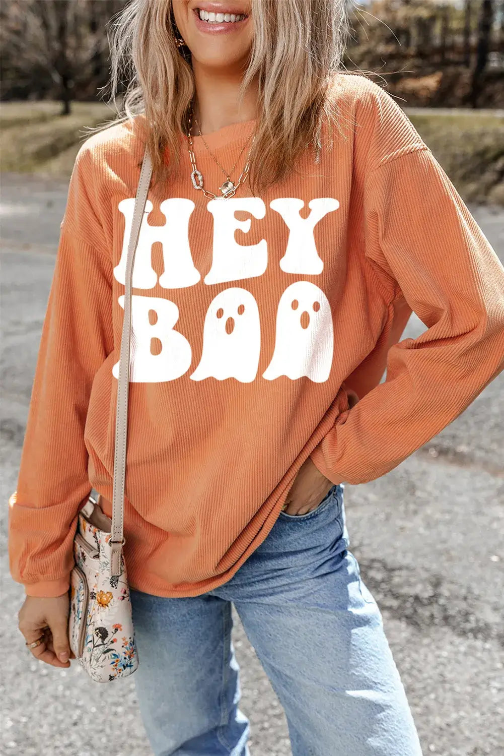 Woman in orange Bella Road Hey Boo Graphic Round Neck Long Sleeve Sweatshirt with blue jeans, accessorized with a crossbody bag and necklace.