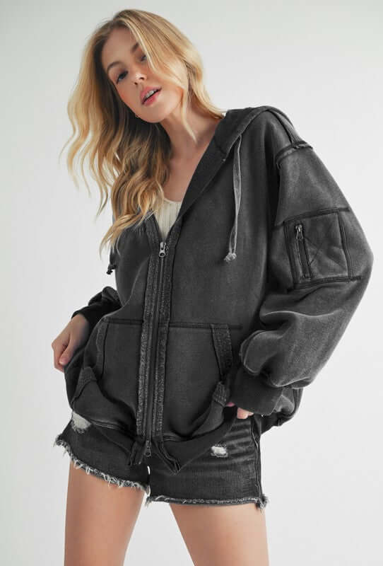 Trendy woman in a black exposed seam zip-up drawstring hooded jacket, styled with denim shorts for a chic look.