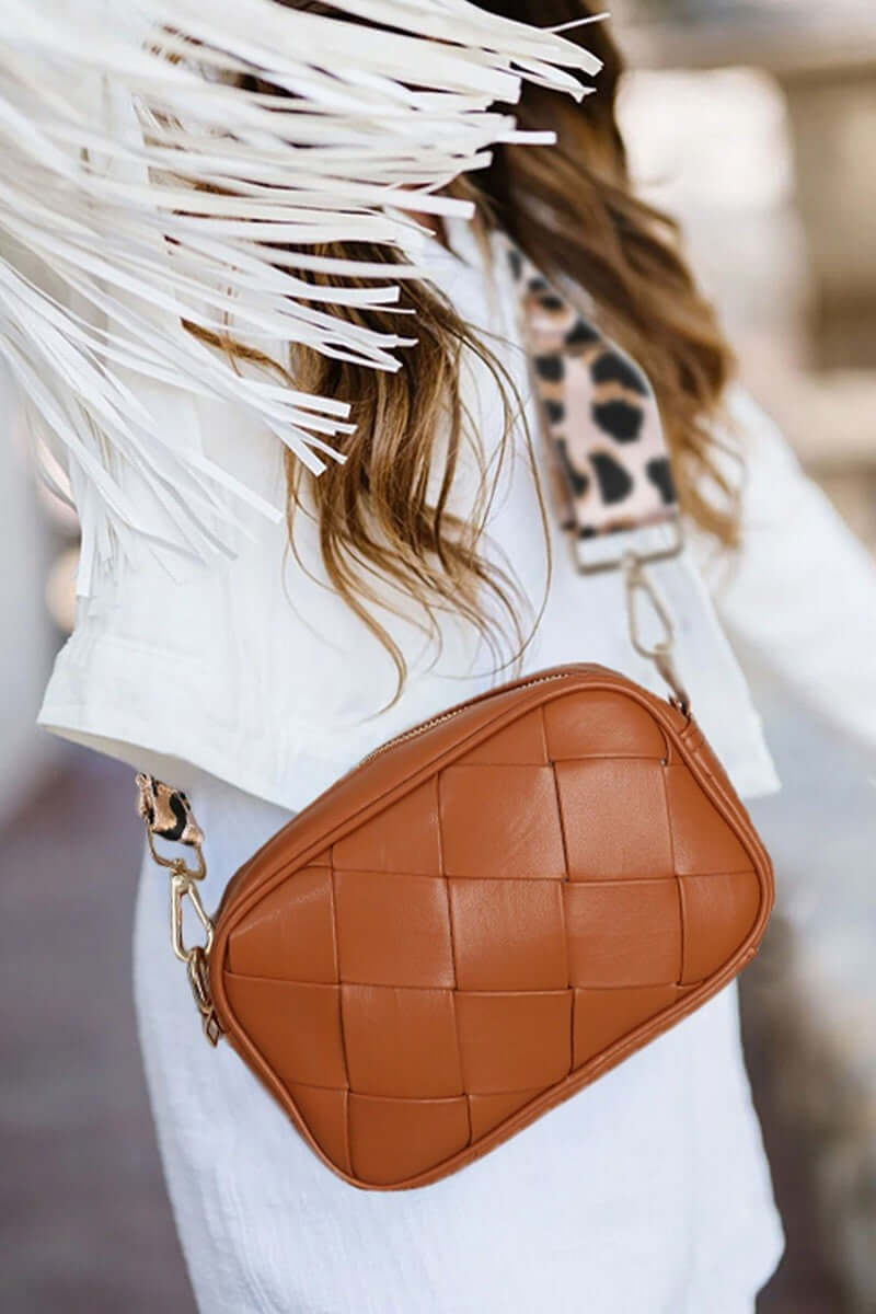 Stylish woman in a white jacket holding a woven tan crossbody bag with leopard strap, showcasing fashion and functionality.