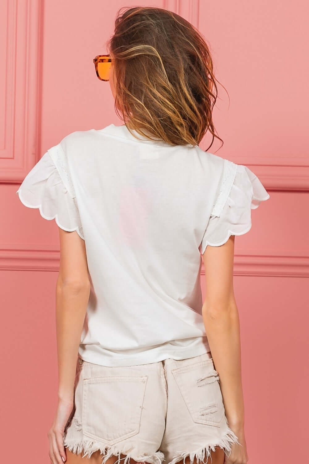 Woman wearing white scallop edged lace trim top with intricate lace detailing, paired with frayed hem shorts, standing against a pink background