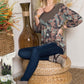 Woman wearing camouflage V-neck balloon sleeve top with dark jeans and suede ankle boots, sitting on a wicker ottoman in a casually decorated room.