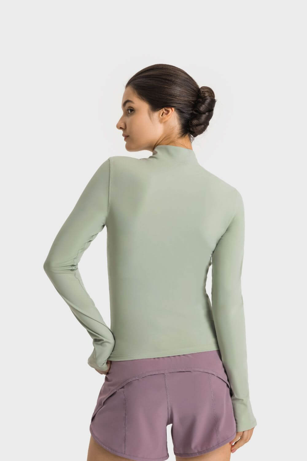 Model showing the back of the Millennia Half Zip Thumbhole Sleeve Sports Top in green, paired with sporty shorts.