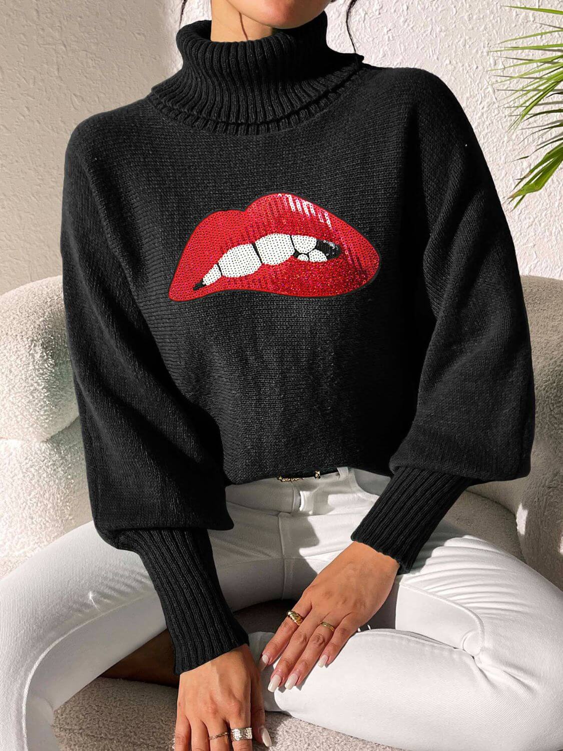 Woman wearing Bella Road Lip Turtleneck Long Sleeve Sweater in black with red lip graphic, sitting on a chair.