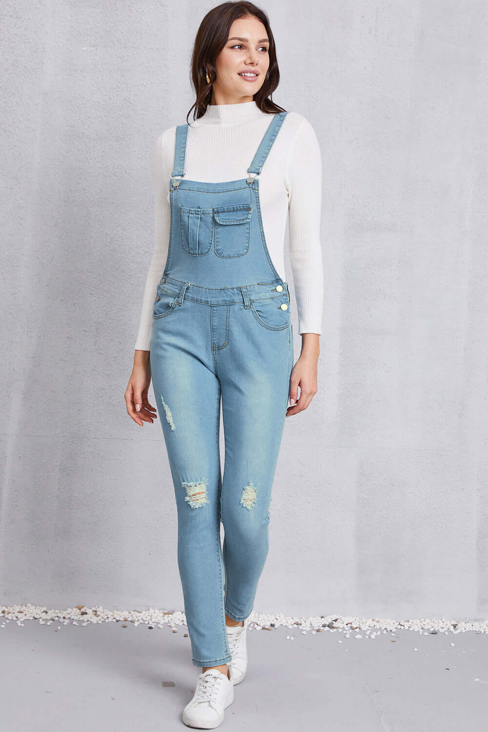 Woman wearing distressed washed denim overalls with pockets, paired with a white long-sleeve top and white sneakers.