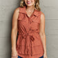 Woman wearing Follow The Light Sleeveless Collared Button Down Top in rust color, paired with blue jeans, standing against a gray wall.