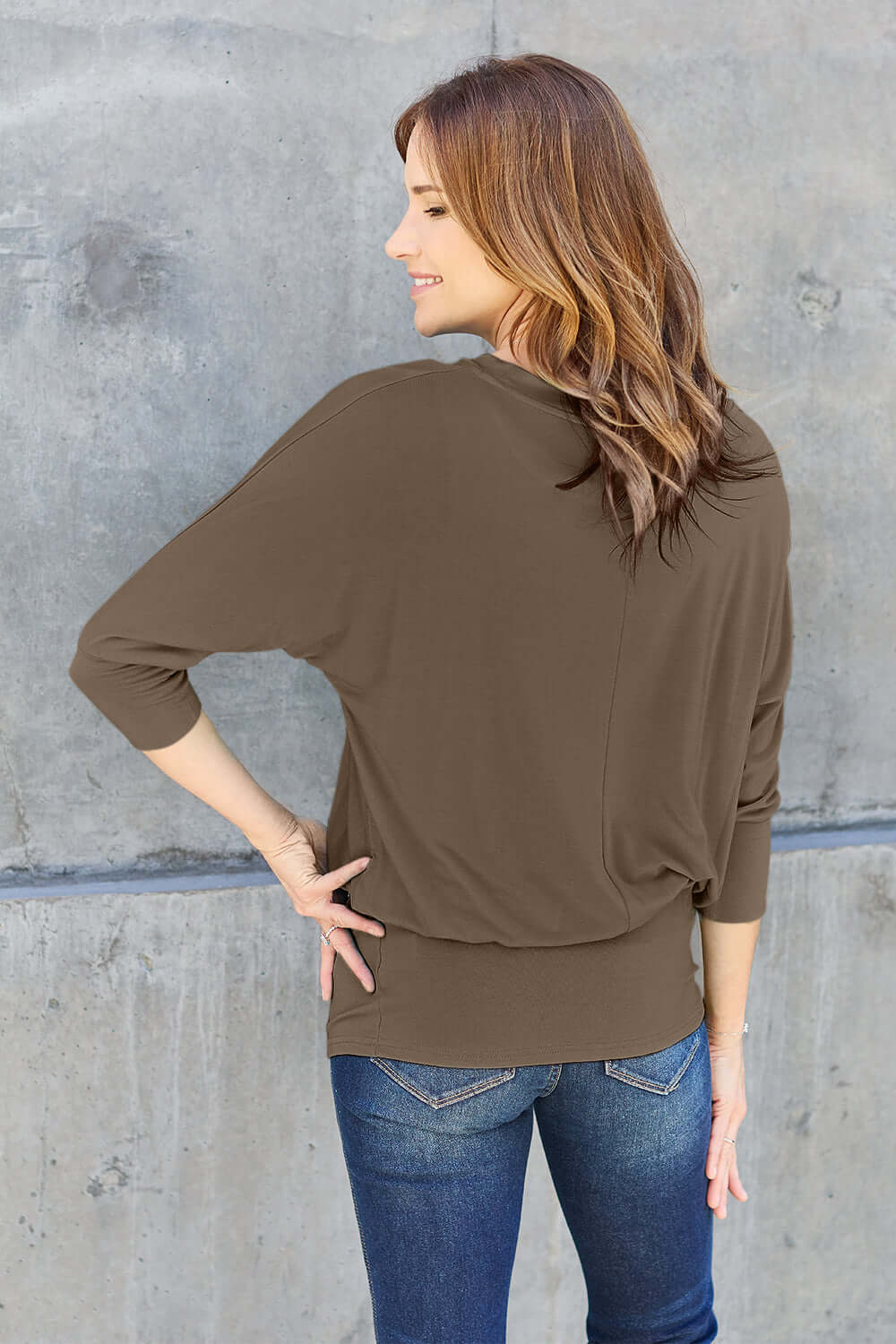 Woman wearing a round neck batwing sleeve top in brown, styled with blue jeans, standing against a concrete wall.