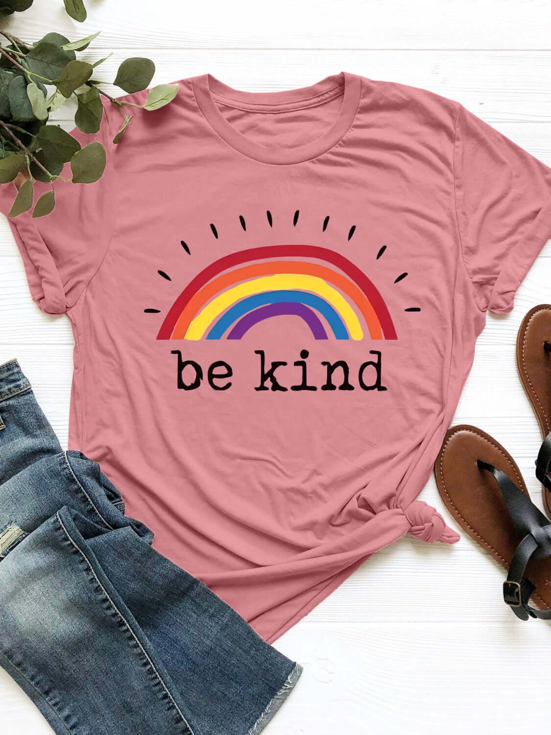 Pink graphic round neck short sleeve T-shirt with "Be Kind" rainbow print, paired with jeans and sandals on a white background.