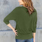 Woman wearing green round neck batwing sleeve top with blue jeans, back view against a concrete wall background.