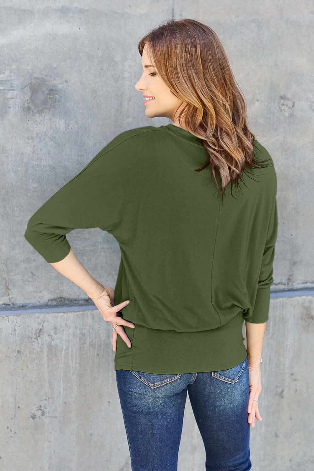 Woman wearing green round neck batwing sleeve top with blue jeans, back view against a concrete wall background.