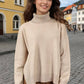 Woman wearing beige turtleneck sweater with side slits and dropped shoulders, standing on a cobblestone street in a charming town.