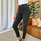 Woman wearing RFM Full Size High Rise Tummy Control Skinny Jeans in black with a white top and black sneakers in a stylish living room.