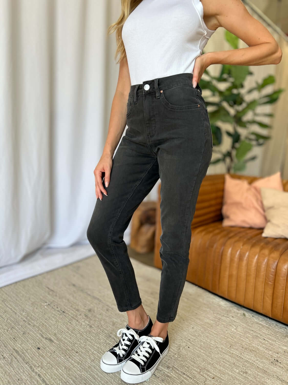 Woman wearing RFM Full Size High Rise Tummy Control Skinny Jeans in black with a white top and black sneakers in a stylish living room.