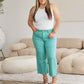 Woman wearing a white sleeveless top and turquoise high-waist crop jeans from RFM Jeans standing in a modern living room.