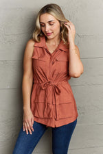 Woman wearing Follow The Light Sleeveless Collared Button Down Top in rust color with tie waist and blue jeans