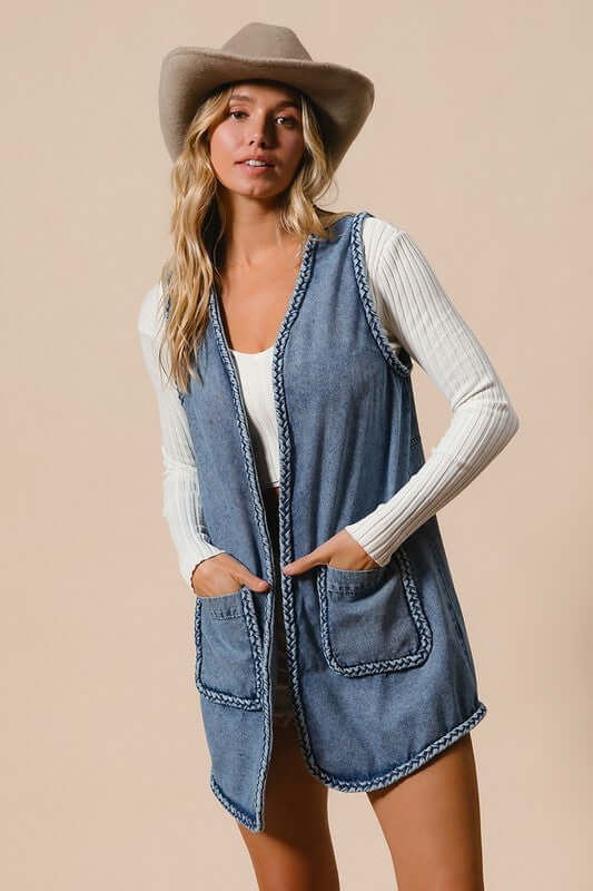 Stylish woman in a BiBi open front denim vest with pockets, paired with a hat against a neutral background.