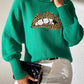 Green Bella Road Lip Turtleneck Sweater with playful design, paired with white pants and clutch.