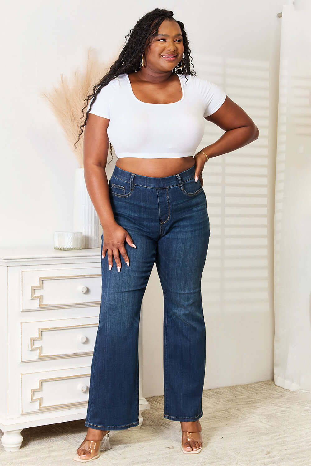 Woman wearing slim bootcut jeans with elastic waistband, standing in a white room, showcasing Judy Blue Jeans.