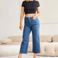 Woman wearing RFM Jeans Crop Chloe high waist tummy control raw hem jeans paired with a black crop top and heels in a stylish living room.