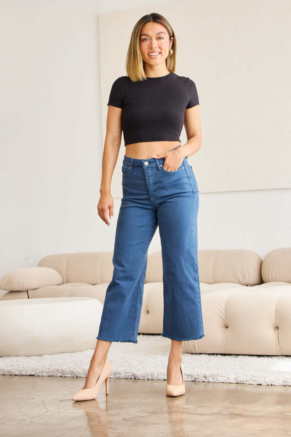 Woman wearing RFM Jeans Crop Chloe high waist tummy control raw hem jeans paired with a black crop top and heels in a stylish living room.