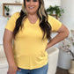 Woman wearing a yellow Bamboo V-Neck High-Low T-Shirt in a living room setting, paired with blue jeans.