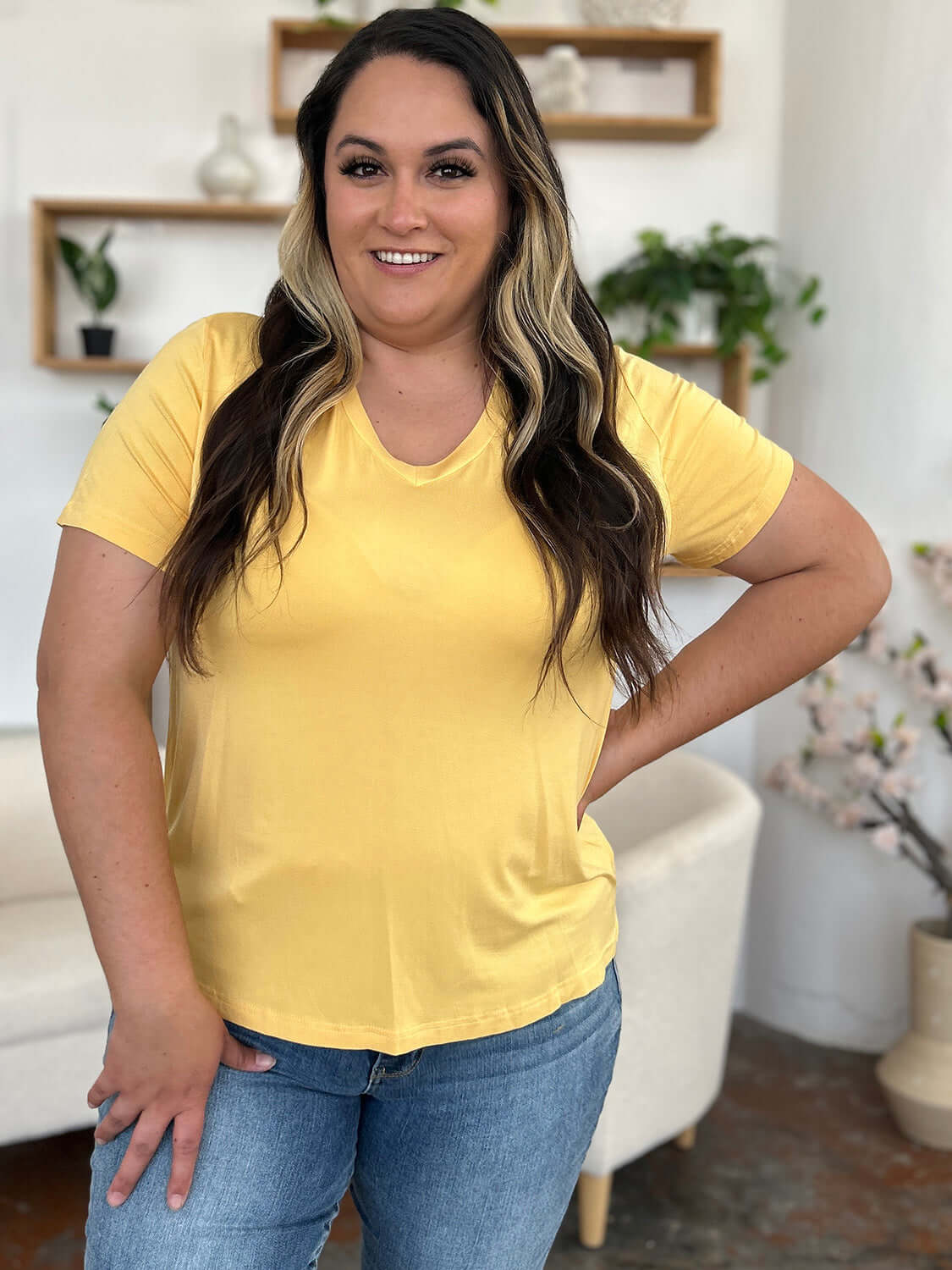 Woman wearing a yellow Bamboo V-Neck High-Low T-Shirt in a living room setting, paired with blue jeans.