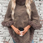 Cozy Bella Road Fuzzy Hem Cable-Knit Poncho in taupe, styled with a coffee cup against a textured brick wall.