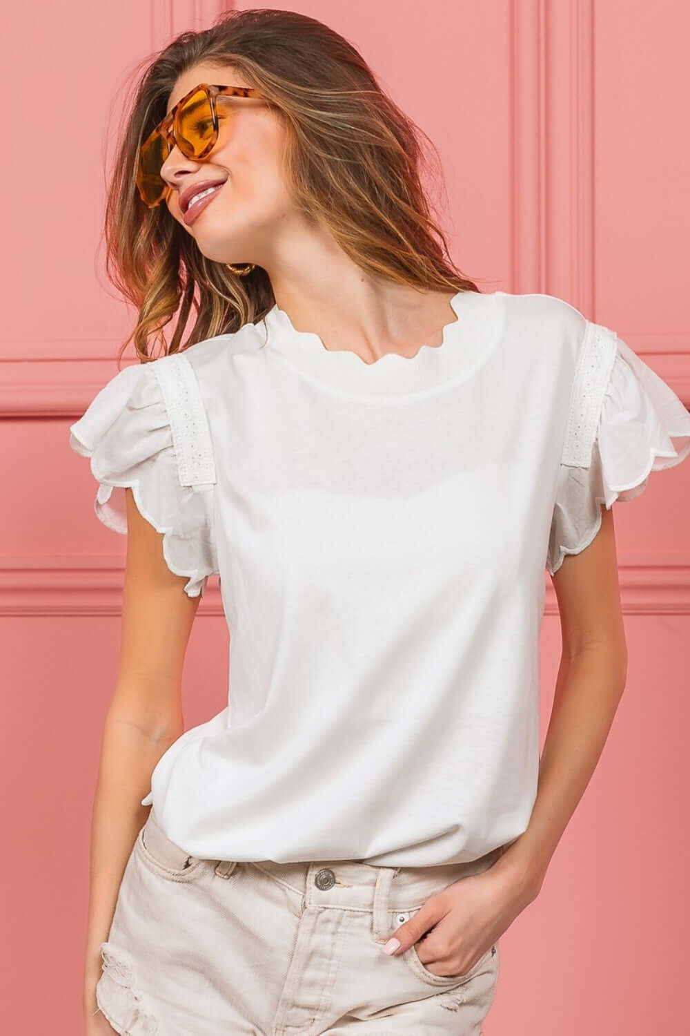 Woman wearing a white scallop edged lace trim top with ruffled sleeves against a pink background, styled with sunglasses and light jeans.