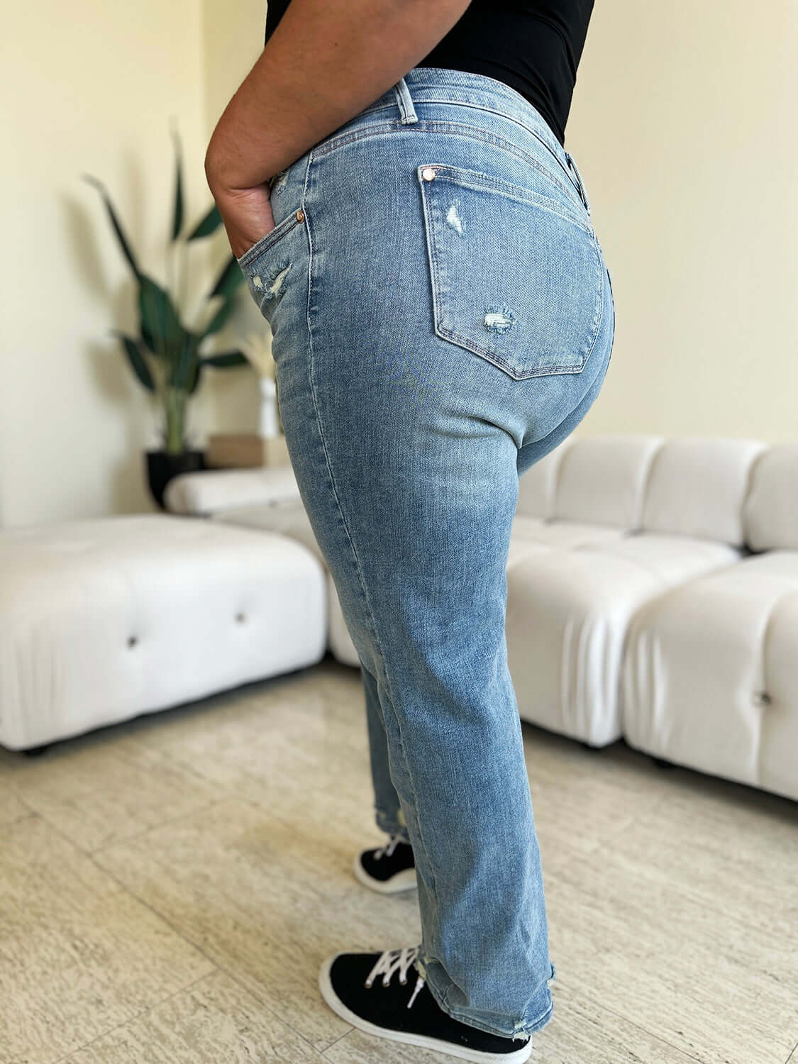 Person wearing high waist distressed straight jeans by Judy Blue, showing back pocket details and straight leg fit, in a stylish living room setting