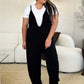 Woman wearing black plunge sleeveless jumpsuit with pockets over white t-shirt standing in stylish living room.