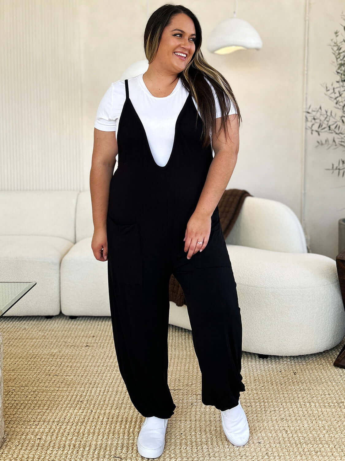 Woman wearing black plunge sleeveless jumpsuit with pockets over white t-shirt standing in stylish living room.