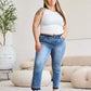 Woman wearing Judy Blue release hem cropped bootcut jeans with a white tank top and sandals in a modern living room.